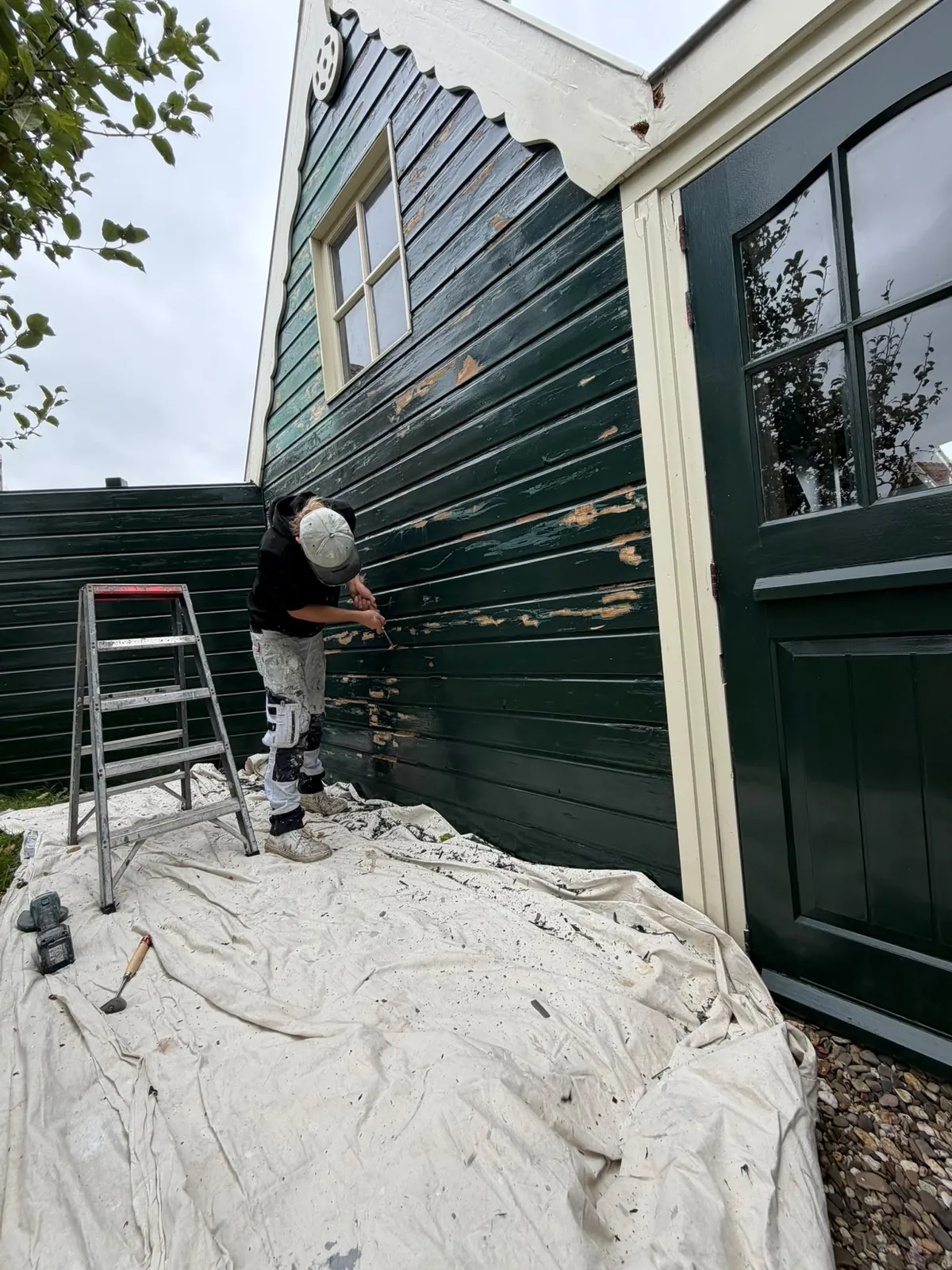 Schilder op een trap bij een donkergroene houten gevel, de grond afgedekt met stoflakens
