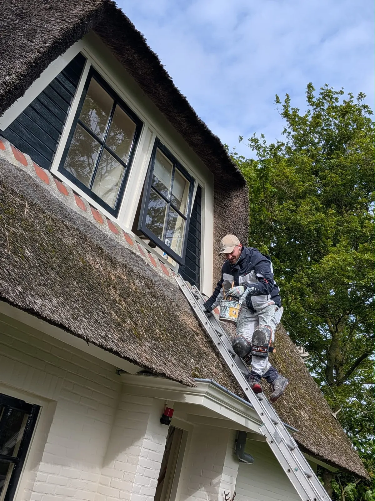 Schilder op een ladder tegen een rieten dak werkt aan de dakkapelkozijnen